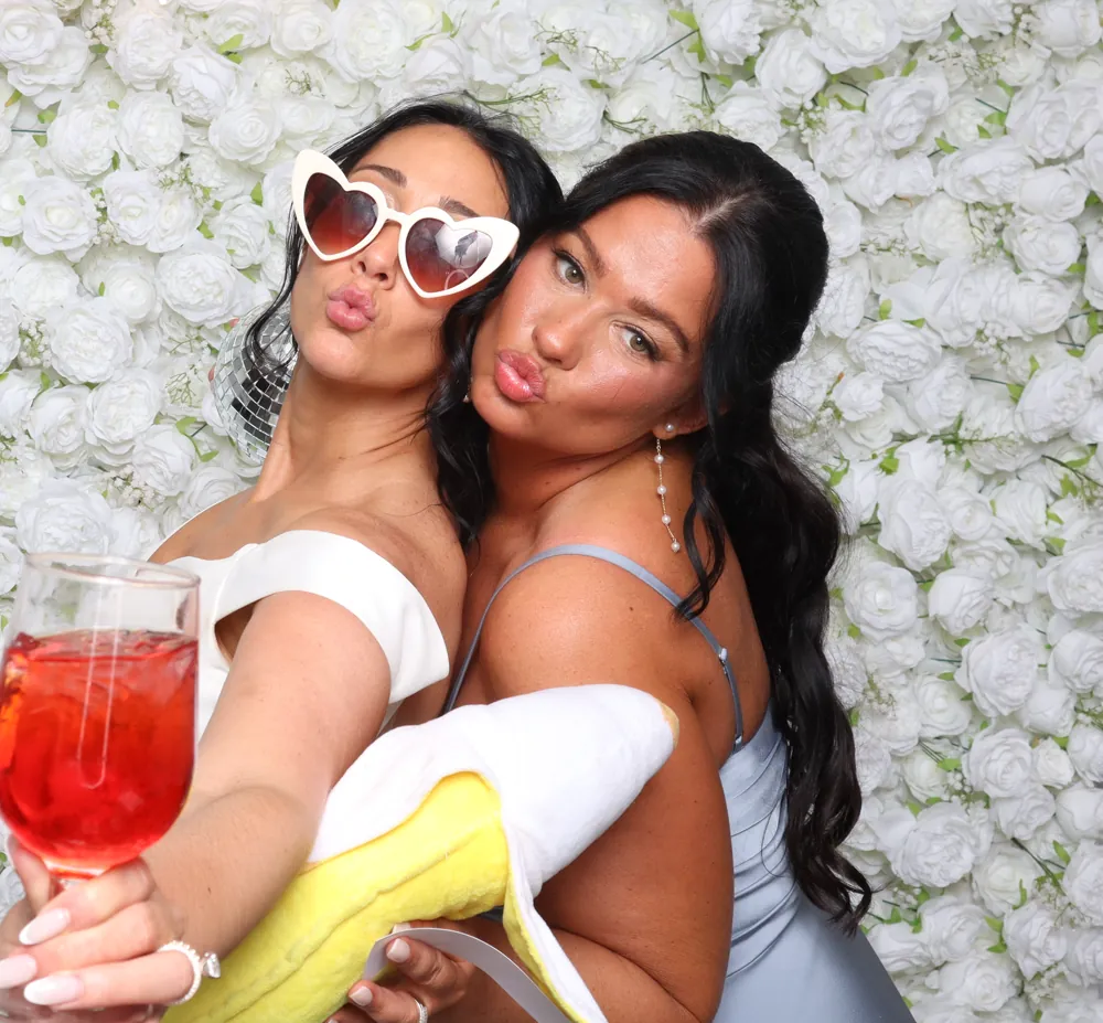 Two guests in heart shaped sunglasses posing against a white flower wall backdrop