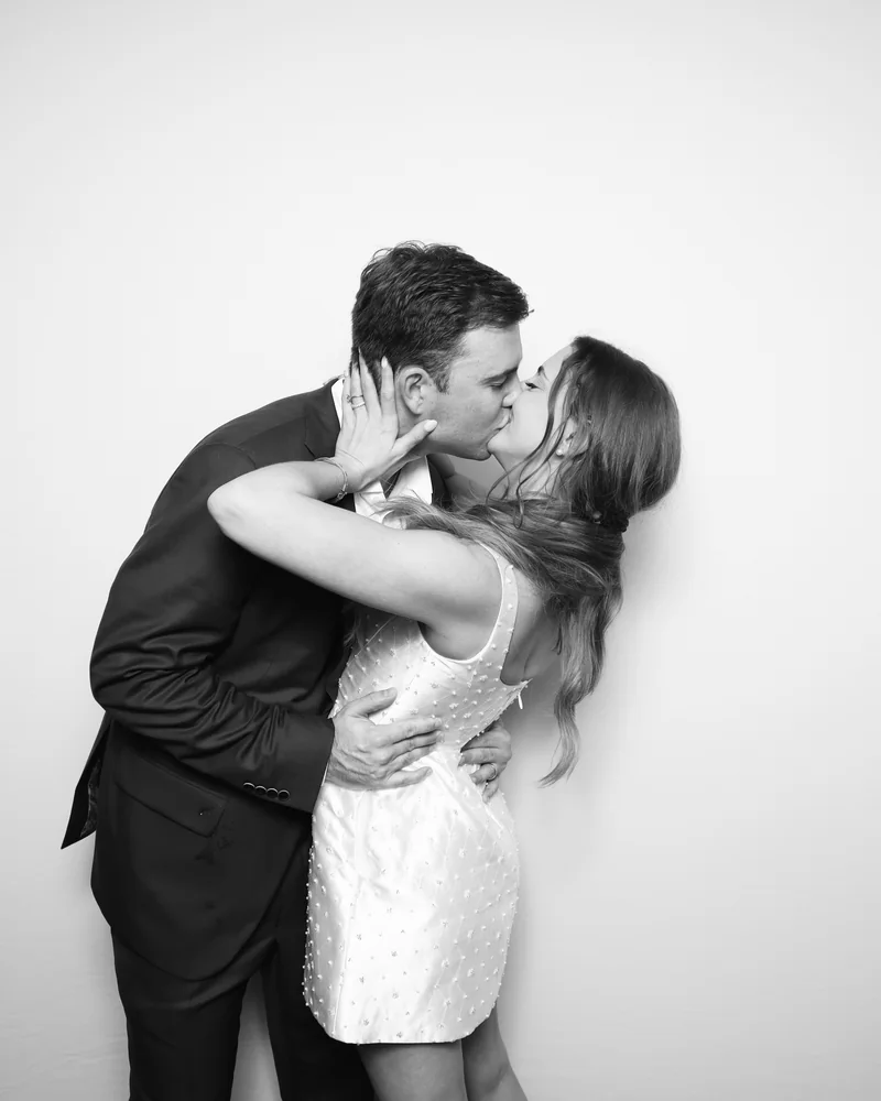 A black and white photo booth portrait of a couple kissing against a white backdrop