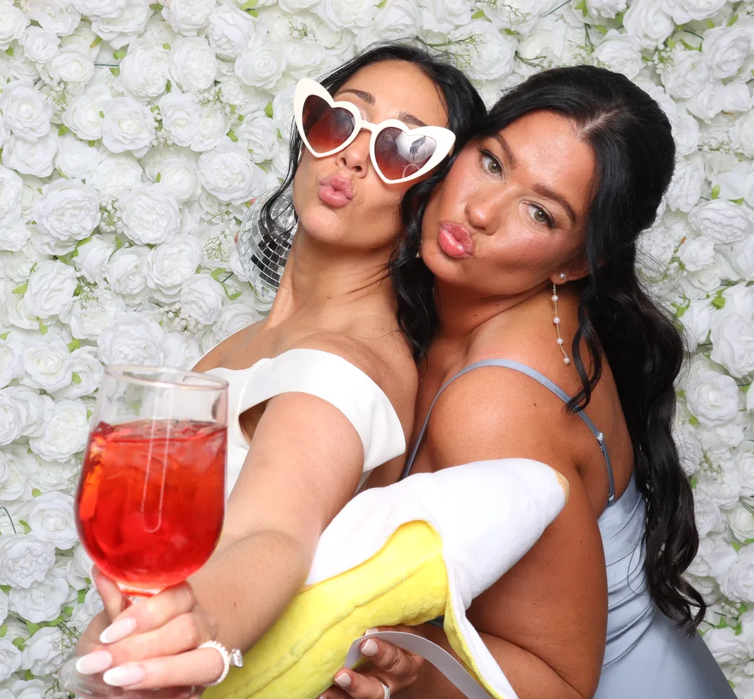 Two wedding guests posing together in front of a white flower wall in the photo booth