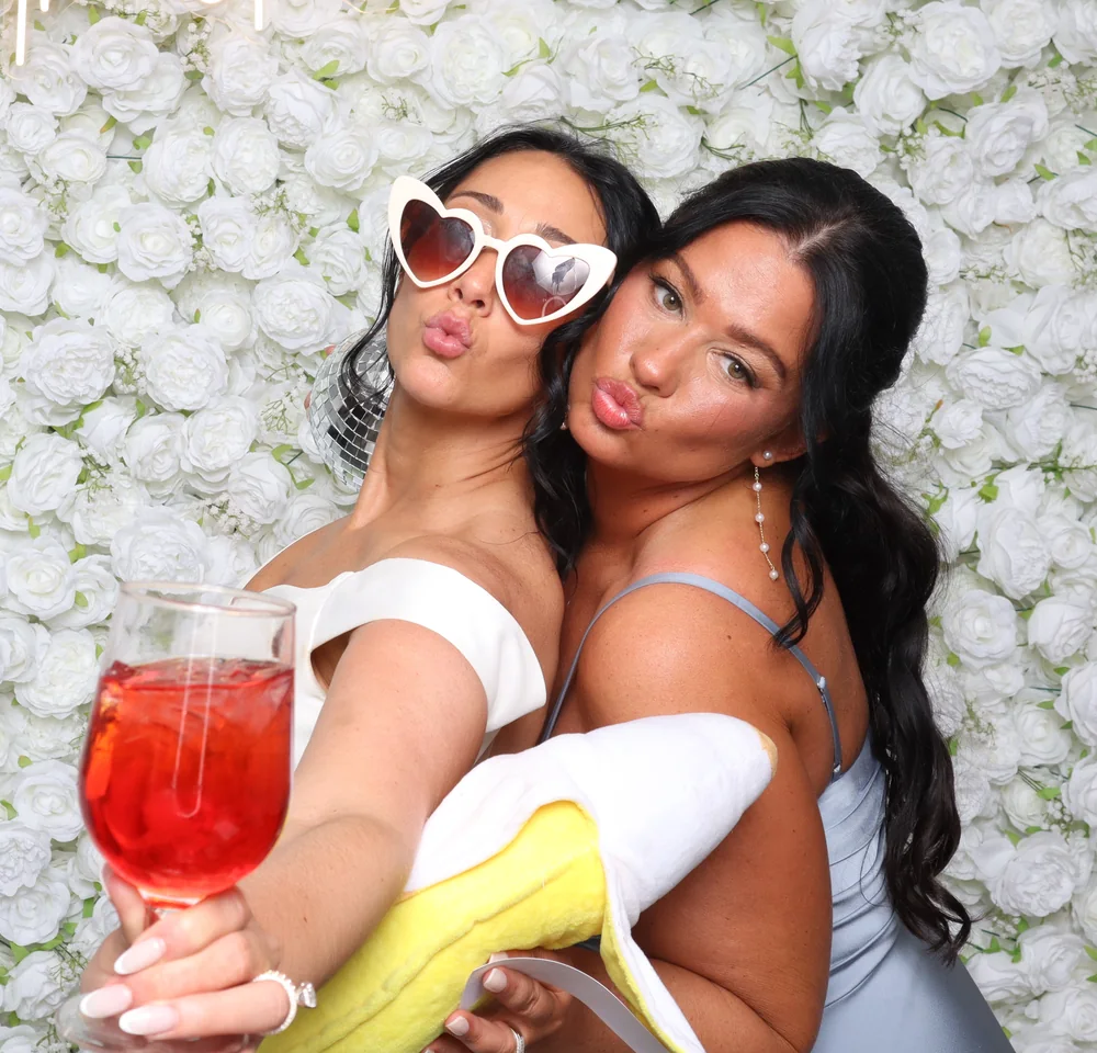 Two guests posing with a drink in front of a white flower wall backdrop