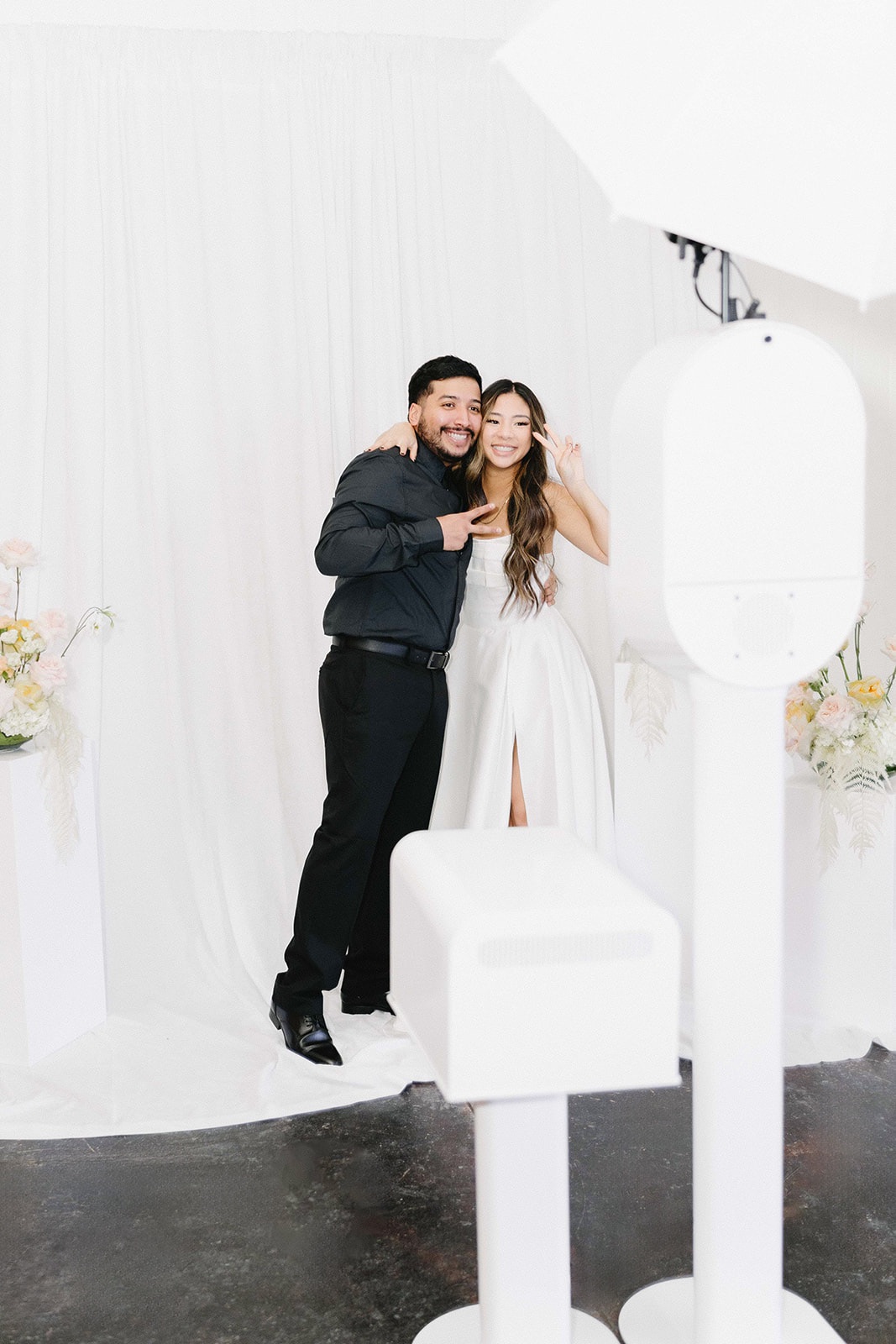 A bride and groom posing together beside the white booth in front of a draped backdrop