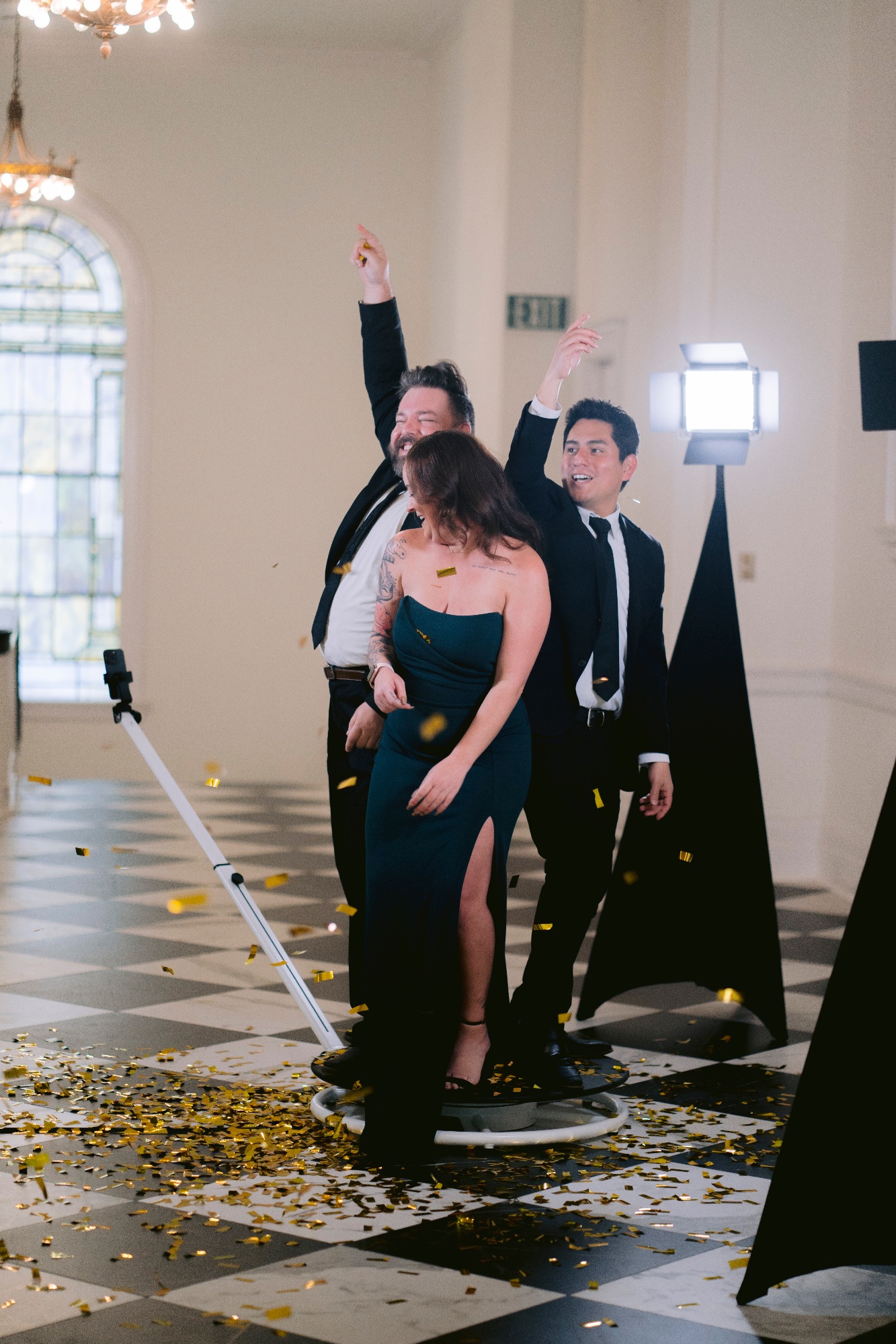Three guests in formalwear cheering on the 360 booth platform as gold confetti falls around them